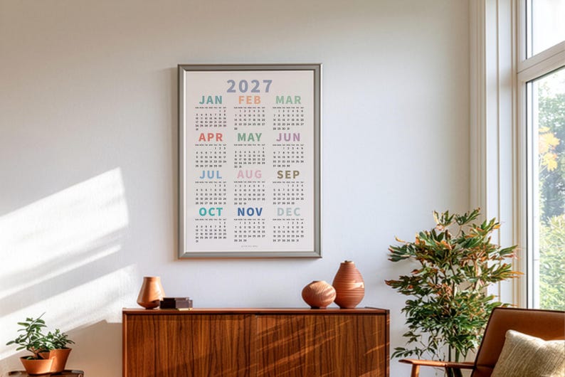 May include: A framed 2027 wall calendar featuring a grid layout of all twelve months. Each month name is printed in a distinct pastel color above its corresponding numerical date grid. The calendar is displayed in a simple silver frame.