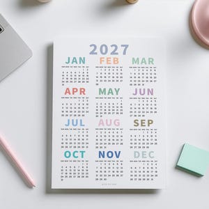 May include: A 2027 wall calendar printed on white paper featuring twelve months arranged in a grid. Each month name is displayed in a distinct pastel color, including blue, orange, green, pink, and purple, with corresponding numerical dates below.