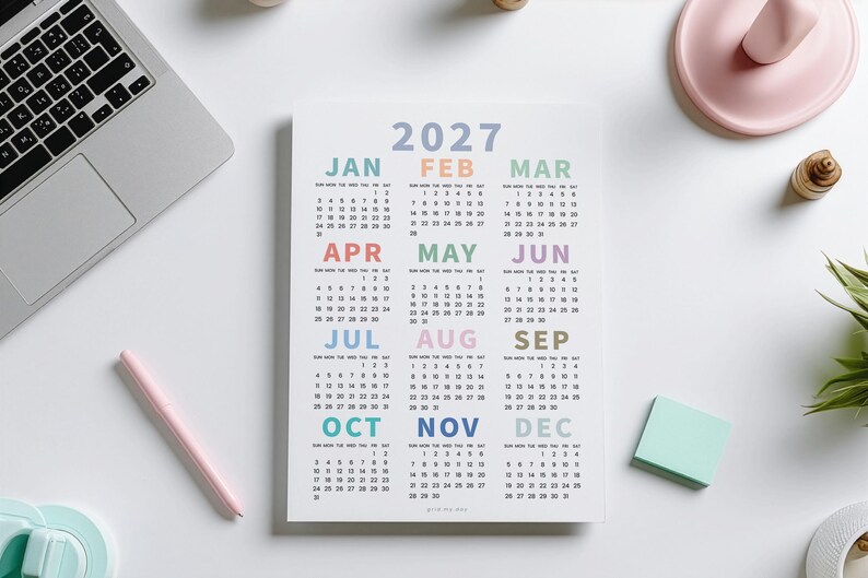 May include: A 2027 wall calendar printed on white paper featuring twelve months arranged in a grid. Each month name is displayed in a distinct pastel color, including blue, orange, green, pink, and purple, with corresponding numerical dates below.