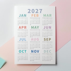 May include: A 2027 wall calendar printed on white paper featuring twelve months arranged in a grid. Each month name is displayed in a different pastel color, including blue, orange, green, pink, purple, and yellow, with black numerical dates.