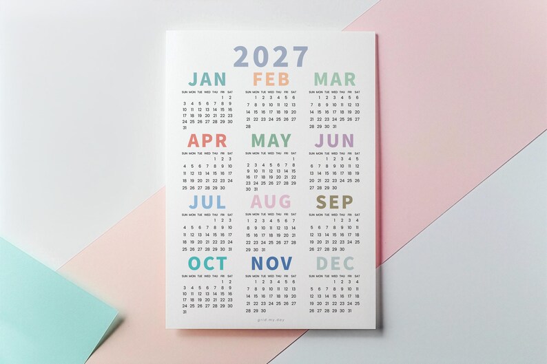 May include: A 2027 wall calendar printed on white paper featuring twelve months arranged in a grid. Each month name is displayed in a different pastel color, including blue, orange, green, pink, purple, and yellow, with black numerical dates.