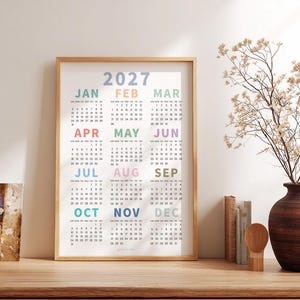 May include: A vertical wall calendar for the year 2027 displayed in a light wood frame. The calendar features twelve months arranged in a grid, with each month name printed in a different pastel color above its respective numerical date grid.