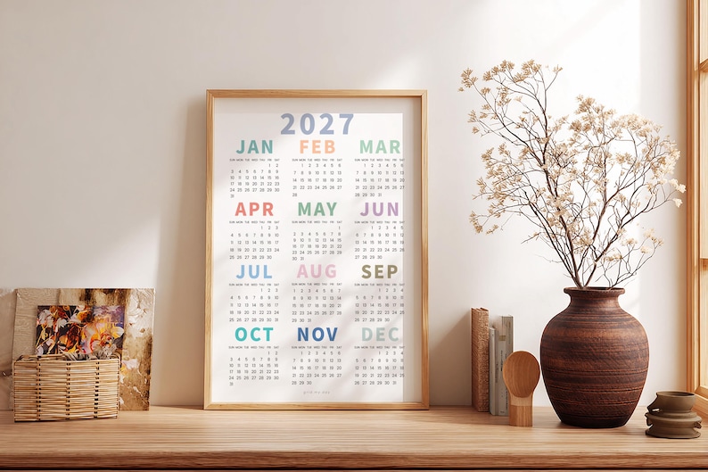 May include: A vertical wall calendar for the year 2027 displayed in a light wood frame. The calendar features twelve months arranged in a grid, with each month name printed in a different pastel color above its respective numerical date grid.