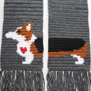 Può includere: Una sciarpa di uncinetto grigia con un'immagine di uncinetto colorata di un cane corgi con un cuore rosso sul petto. La sciarpa ha frange grigie su entrambe le estremità.