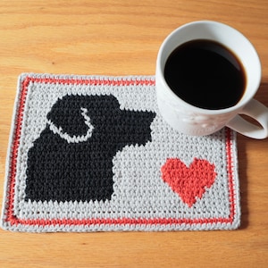 May include: A crocheted coaster with a black dog silhouette and a red heart on a grey background. The coaster has a red border and is next to a white mug filled with coffee. A delightful item for the home.