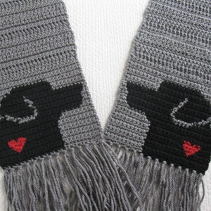 Könnte beinhalten: Grauer Häkel-Schal mit schwarzen Hundesilhouetten und roten Herzen. Der Schal hat Fransen an den Enden.