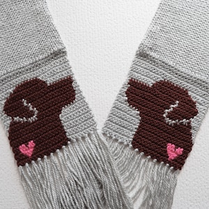 May include: A gray knitted scarf with brown dog silhouettes and pink hearts. The scarf has fringe on the ends.