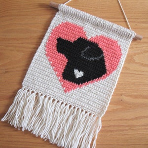 May include: A crocheted wall hanging featuring a pink heart with a black silhouette of a dog inside. The hanging has a fringe at the bottom.
