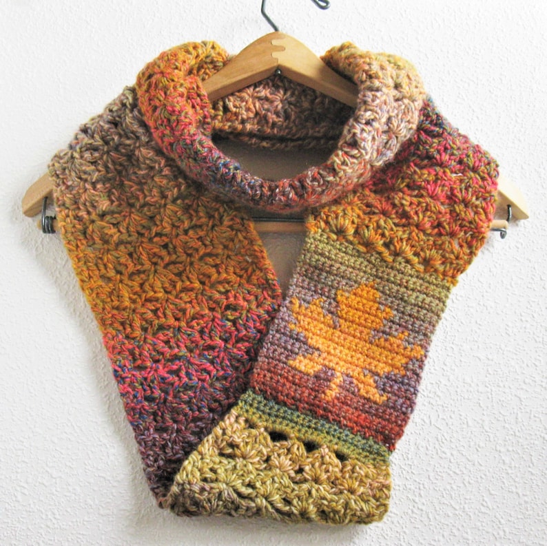 May include: A handmade crochet cowl scarf in autumn colors of orange, red, and brown, hanging on a wooden hanger. The scarf features a leaf design and a textured stitch pattern, perfect for fall and winter wear.