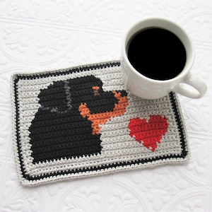 May include: Crocheted coaster featuring a black and tan dog silhouette with a red heart. The coaster is on a white surface with a cup of coffee.