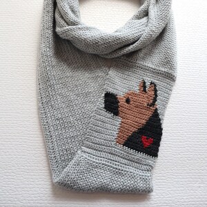 May include: A gray knitted scarf featuring a dog design. The dog's head is brown with a black nose and ears, and a black body with a red heart. The scarf is made of a soft, knitted material.