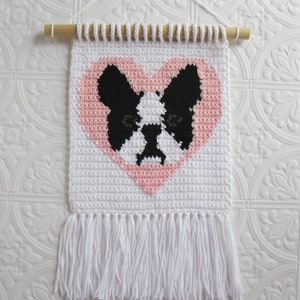 May include: White crocheted wall hanging with a pink heart featuring a black and white French bulldog design. The hanging has a white fringe.