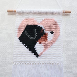 May include: A white crocheted wall hanging with a pink heart in the center. The heart is made up of two dog faces, one black and one brown. The wall hanging has a wooden dowel at the top and white fringe at the bottom.