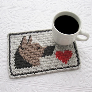 May include: A crocheted coaster featuring a brown dog silhouette and a red heart. The coaster is on a white surface with a cup of coffee.