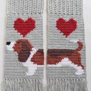 May include: A gray crocheted scarf with a brown and white dog and two red hearts. The scarf has fringe on the bottom.