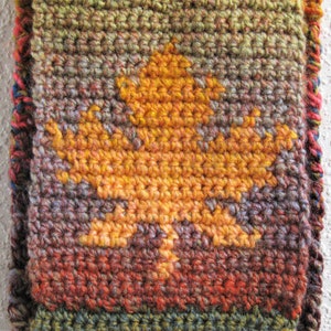 May include: A handmade crochet scarf featuring a large, golden-yellow leaf design. The scarf has a textured appearance with a mix of autumnal colors, including shades of orange, brown, green, and red. The edges are finished with a colorful border.