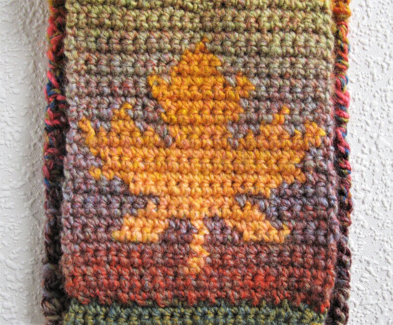 May include: A handmade crochet scarf featuring a large, golden-yellow leaf design. The scarf has a textured appearance with a mix of autumnal colors, including shades of orange, brown, green, and red. The edges are finished with a colorful border.