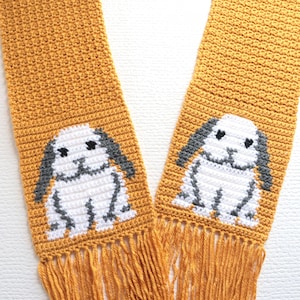 May include: A mustard yellow crochet scarf featuring two identical white and grey lop-eared rabbit motifs. The scarf is constructed with a dense stitch pattern and finished with long, matching mustard yellow fringe at both ends.