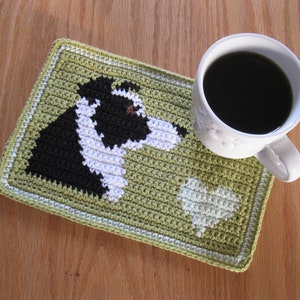 May include: Green crocheted coaster featuring a white and black dog design and a green heart. A white coffee mug with black coffee is placed on top of the coaster.