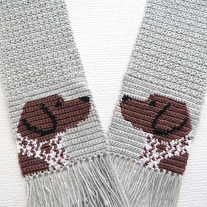 以下が含まれることがあります： グレーのクロシェ編みスカーフで、茶色の犬の顔が描かれ、両端に白と茶色のスカーフが付いています。スカーフは両端にフリンジが付いています。