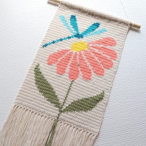 May include: A cream-coloured, crocheted wall hanging with a pink daisy, yellow centre, and blue dragonfly design. Green leaves and a fringe adorn the bottom. A wooden dowel is at the top for hanging. Measurements are in centimetres.