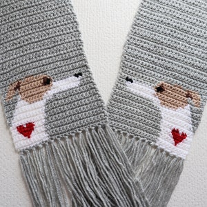 May include: A handmade gray crochet scarf featuring a dog design. The dog has a white body, tan ears, and a red heart. The scarf has fringe at the ends. The scarf is approximately 60 inches long.
