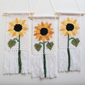 May include: Three white crocheted wall hangings with a sunflower design. Each hanging features a yellow sunflower with a brown center and green leaves. The hangings have white fringe at the bottom.