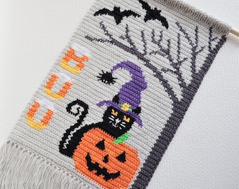 May include: A crocheted Halloween wall hanging with a black cat wearing a purple witch hat and holding a jack-o'-lantern. The hanging has a fringe border and the word "BOO" is crocheted in orange and yellow yarn.