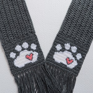 May include: Gray crocheted scarf with white paw prints and pink hearts. The scarf has fringe on the ends.