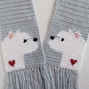 May include: A grey crocheted scarf with white and red crocheted designs of two cartoon bears with hearts. The scarf has grey fringe on the ends.