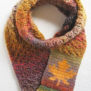 May include: A handmade, crocheted infinity scarf in autumn colors. The scarf features a repeating pattern of orange, red, and yellow yarn. A section of the scarf displays a yellow leaf design. The scarf is a cozy accessory for fall.