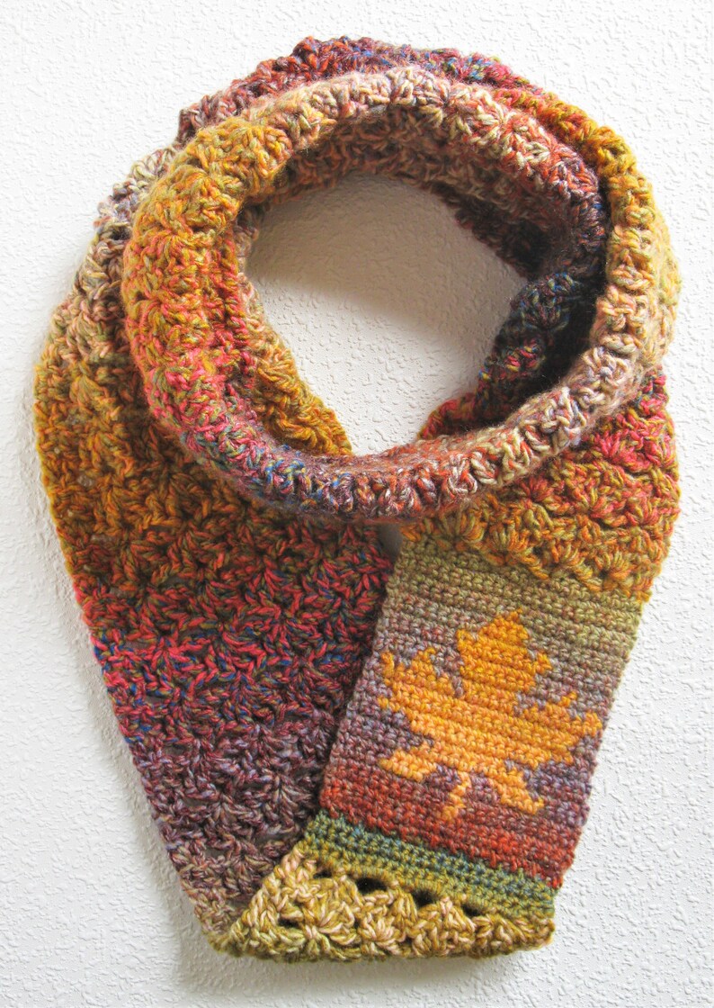 May include: A handmade, crocheted infinity scarf in autumn colors. The scarf features a repeating pattern of orange, red, and yellow yarn. A section of the scarf displays a yellow leaf design. The scarf is a cozy accessory for fall.