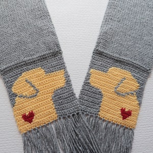 May include: Gray knitted scarf with yellow dog silhouettes and red hearts. The scarf has fringe on the bottom.
