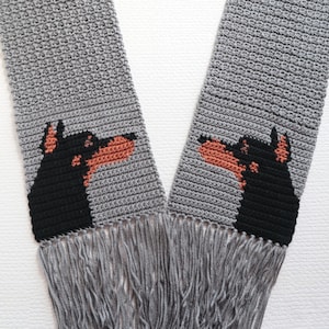 May include: A gray crochet scarf featuring a repeating pattern of a black and tan Doberman Pinscher dog head. The scarf has a fringe at the bottom. The dog's head is on both ends of the scarf.