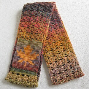 May include: A handmade crochet scarf in autumnal colors, featuring a gradient of orange, red, and brown. The scarf has a leaf design in the center and openwork detailing. The scarf is approximately 60 inches long.