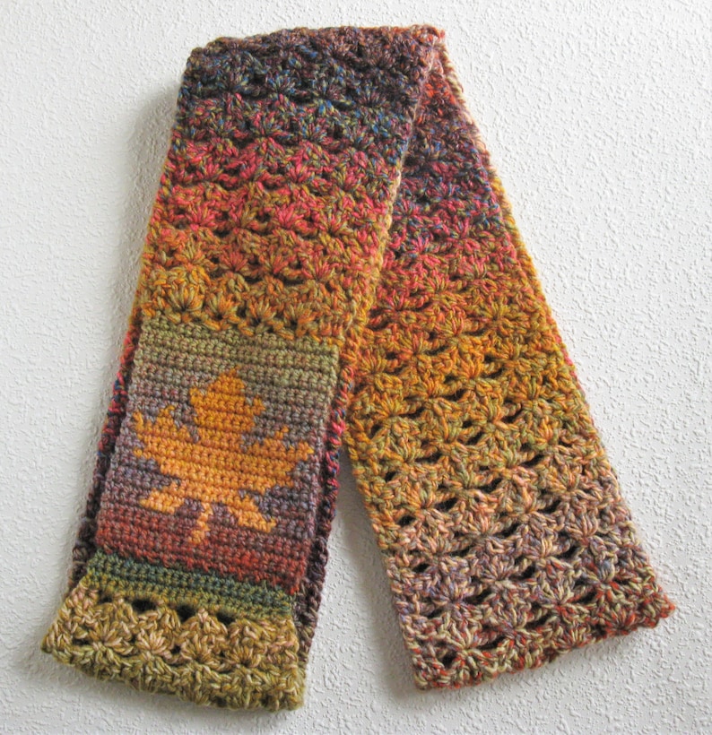May include: A handmade crochet scarf in autumnal colors, featuring a gradient of orange, red, and brown. The scarf has a leaf design in the center and openwork detailing. The scarf is approximately 60 inches long.