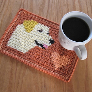 May include: A crocheted coaster featuring a dog design in shades of white, yellow, and pink. The coaster is placed on a wooden surface with a white coffee mug on top.