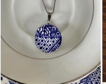 May include: A silver necklace with a round pendant featuring a blue and white patterned design. The pendant is suspended from a delicate chain and rests on a white plate.