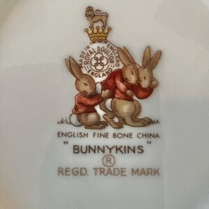 May include: A white English fine bone china plate with a Bunnykins design. The plate features two brown bunnies wearing red jackets, with the words "Bunnykins" and "Regd. Trade Mark" printed below.