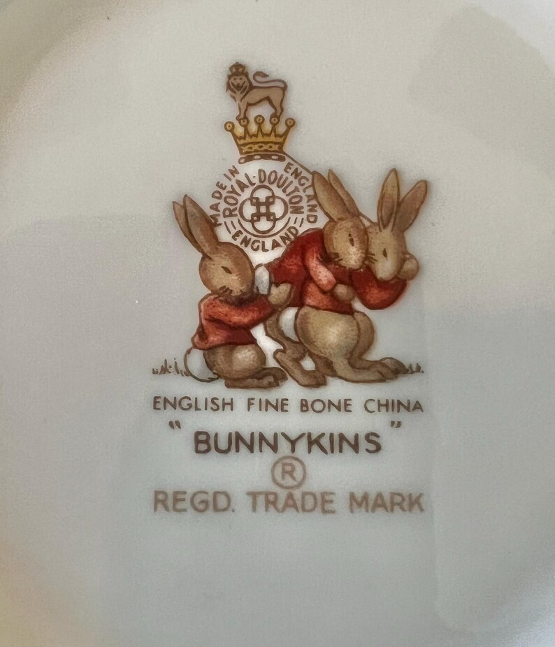 May include: A white English fine bone china plate with a Bunnykins design. The plate features two brown bunnies wearing red jackets, with the words "Bunnykins" and "Regd. Trade Mark" printed below.