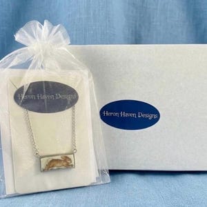 May include: A silver-toned necklace with a rectangular pendant featuring a brown rabbit design, presented in a sheer gift bag. The bag is tied with a white ribbon and includes a card with the text "Heron Haven Designs". A white box with the same logo is in the background.