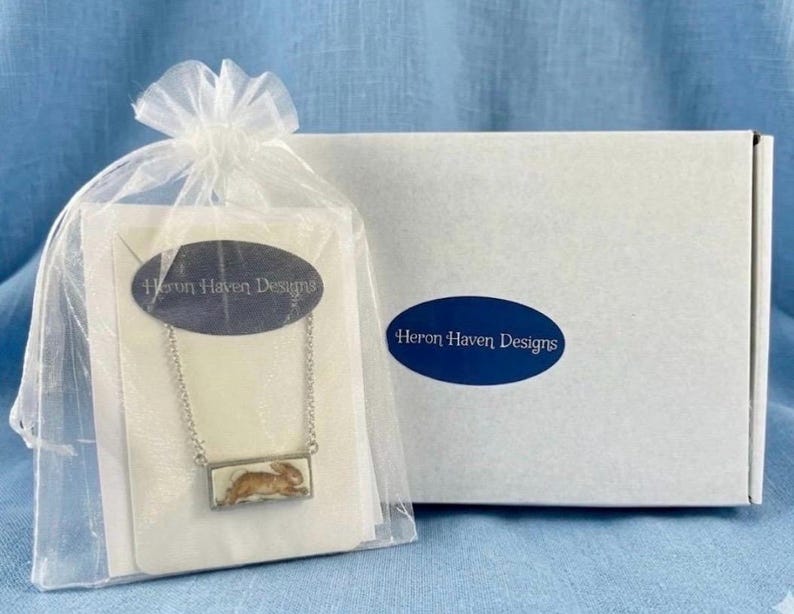 May include: A silver-toned necklace with a rectangular pendant featuring a brown rabbit design, presented in a sheer gift bag. The bag is tied with a white ribbon and includes a card with the text "Heron Haven Designs". A white box with the same logo is in the background.