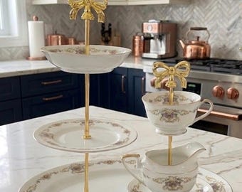 May include: A three-tiered serving stand with gold-colored accents and a bow-shaped handle. The stand holds white porcelain plates and bowls with floral patterns. A teacup and creamer set are also displayed, creating an elegant presentation.