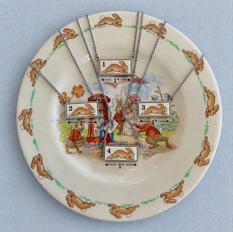 May include: Four silver-tone necklaces with rectangular pendants, each featuring a brown rabbit design. The pendants are approximately 1 inch long. The necklaces are displayed on a decorative plate with a rabbit and floral pattern.