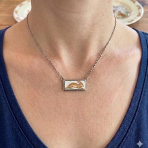 May include: A silver-toned necklace featuring a rectangular pendant with a painted image of a brown rabbit. The rabbit is depicted in a running pose against a white background. The necklace is worn on a person.