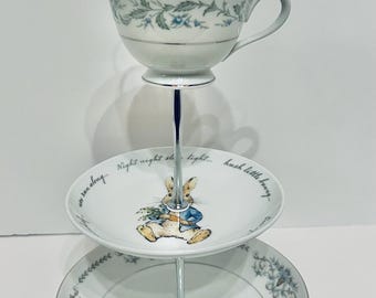 May include: A three-tiered serving stand with a silver handle. The top tier holds a teacup with floral designs. The middle tier features a plate with a Peter Rabbit illustration and text. The bottom tier is a plate with floral designs.