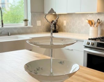May include: A three-tiered serving stand with a silver handle and support. The white ceramic plates have a floral design and a silver rim. The stand is on a wooden surface in a kitchen setting.
