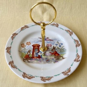 May include: A two-tiered serving tray with a gold-colored handle. The white plates feature a bunny illustration with a border of brown bunnies. The center of the plate has a colorful scene with bunnies and a mailbox.