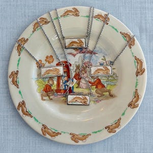 May include: Six silver-toned necklaces with rectangular pendants featuring a brown rabbit design. The necklaces are displayed on a decorative plate with a rabbit and storybook illustration. The plate has a green border and a light blue background.
