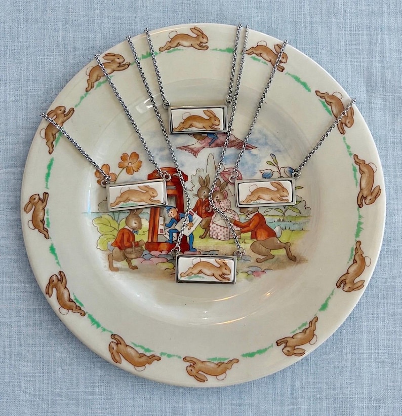 May include: Six silver-toned necklaces with rectangular pendants featuring a brown rabbit design. The necklaces are displayed on a decorative plate with a rabbit and storybook illustration. The plate has a green border and a light blue background.